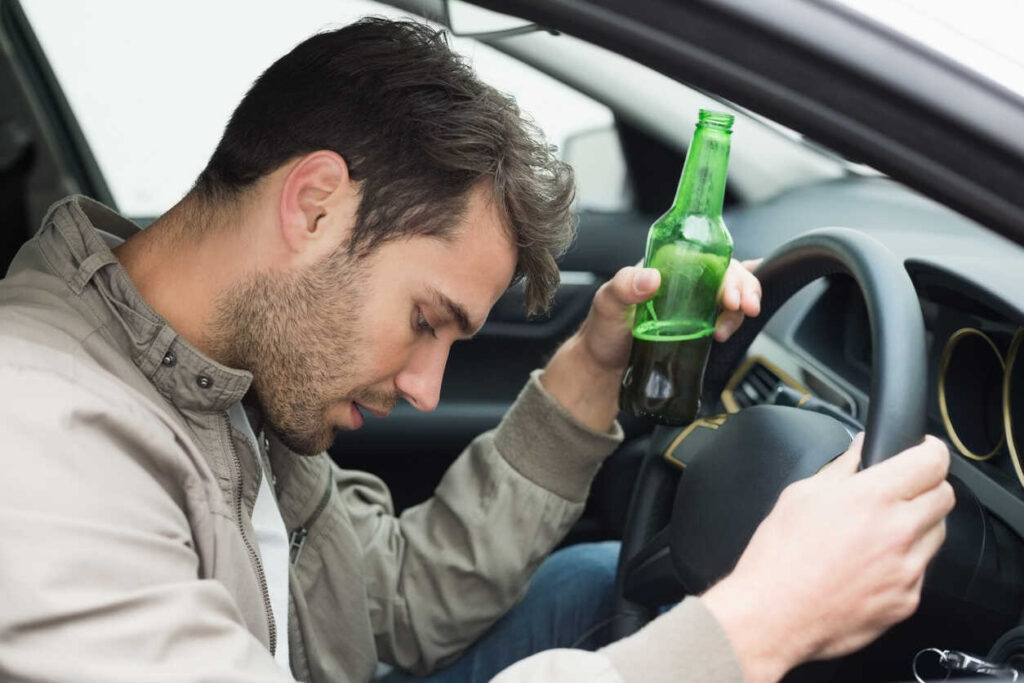 A drunk driver holding a green beer bottle while sitting in the driver's seat, reflecting the reasons why drunk drivers often suffer less harm than their victims in crashes.