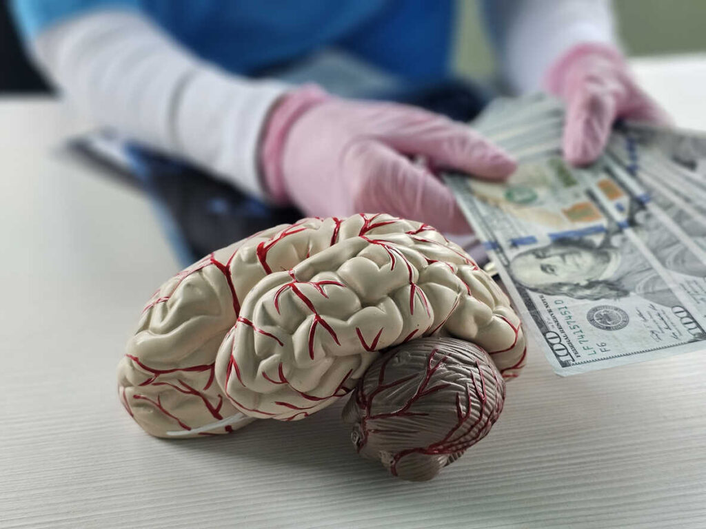 A model of a human brain sits beside a stack of hundred-dollar bills held by gloved hands, symbolizing the high cost of brain injury recovery and the financial assistance a lawyer can help secure.