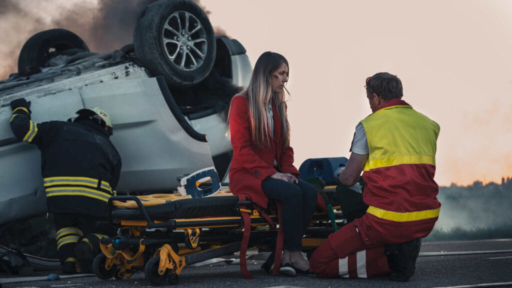 A distressed woman sits on an ambulance stretcher while a paramedic assists her, with an overturned car and firefighter in the background, illustrating a passenger injury accident and the legal rights under New Hampshire and Massachusetts law.
